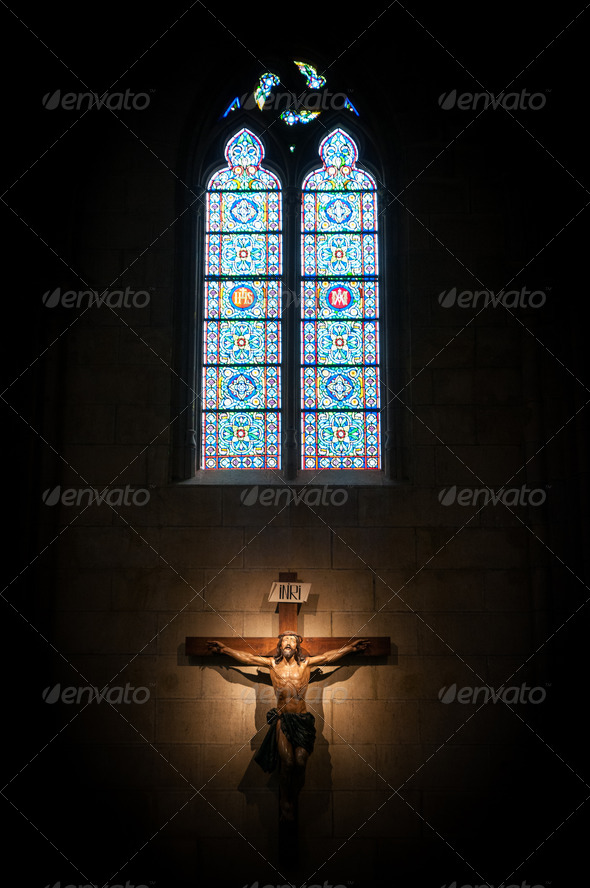 Crucifix on wall in spotlight inside old dark church or cathedral. Jesus Christ on cross on contrast with big bright stained glass window. Religion, belief and hope. Holy and sacred places.