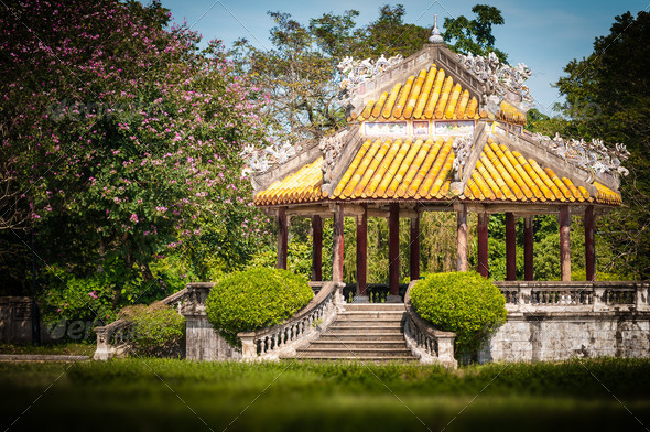Beautiful architecture in traditional asian style. Stairs to ancient building with golden roof and ornate details. Big tree with pink flowers, green lawn. Bright summer day, tranquil landscape.