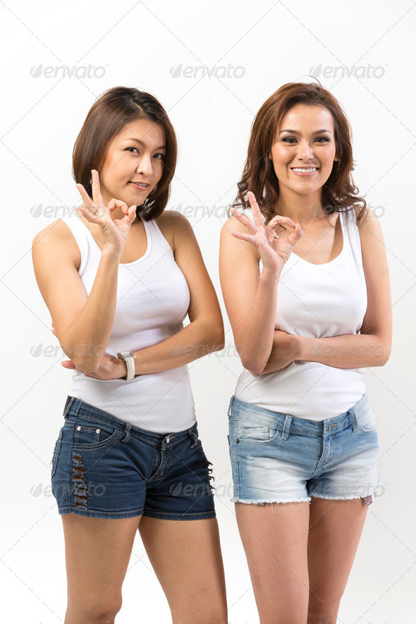 Portrait of two happy Asian Women doing OK symbol. Young fresh Chinese female model Standing in front of a white wall.