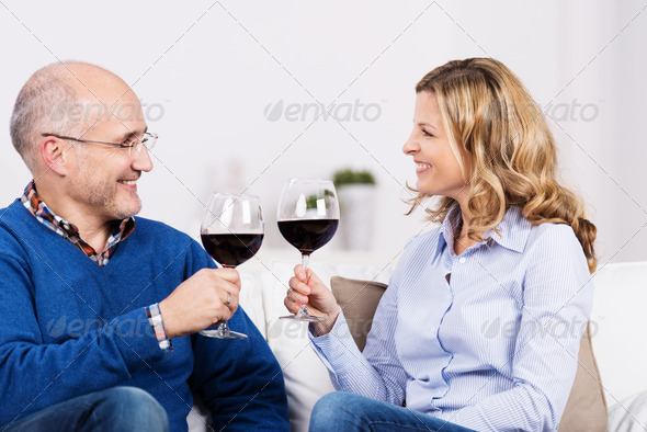 Attractive couple celebrating over a glass of wine sitting in the living room looking at each other with happy smiles as they raise their glasses in a toast