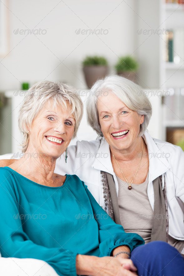 Two happy laughing senior women friends sitting on a sofa in the living room smiling at the camera with optimism
