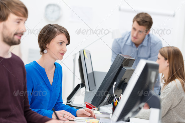 Competent businesswoman working in a busy office sitting smiling at her desktop computer surrounded by hardworking colleagues