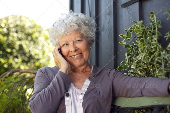 Happy elderly woman sitting on a bench in backyard talking on mobile phone and smiling